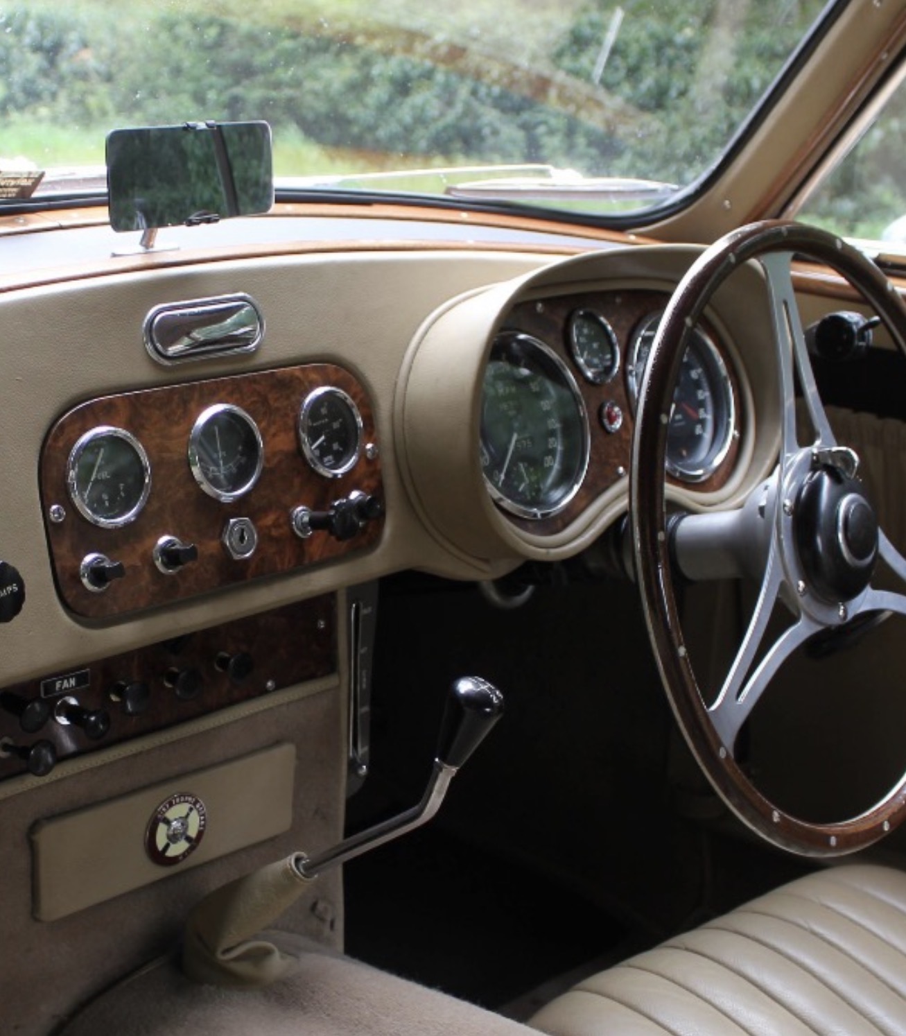 The right-hand-drive cockpit of an AC Aceca, with a burr walnut instrument panel carrying Smiths gauges, a wood-rimmed four-spoke steering wheel, a chrome gear lever and cream leather trim.