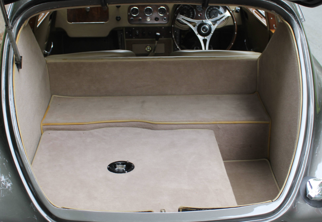 The luggage area of an AC Aceca seen square-on through the open tailgate, fully lined in beige carpet with yellow piping, a stepped floor and a chrome filler cap set into the deck.