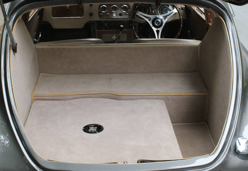 The luggage area of an AC Aceca seen square-on through the open tailgate, fully lined in beige carpet with yellow piping, a stepped floor and a chrome filler cap set into the deck.