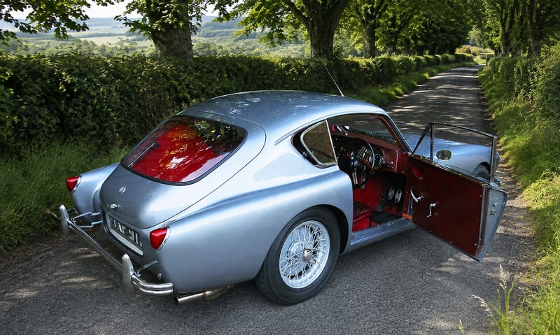 Silver AC Aceca coupé with red interior and door open, rear three-quarter view on a country lane