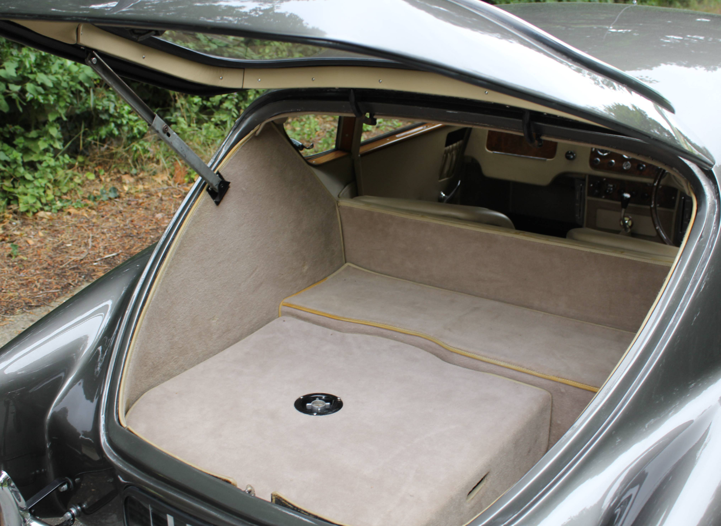 The rear of a grey AC Aceca with the one-piece tailgate raised on its stay, looking down into the carpeted luggage compartment and through to the seats and dashboard.