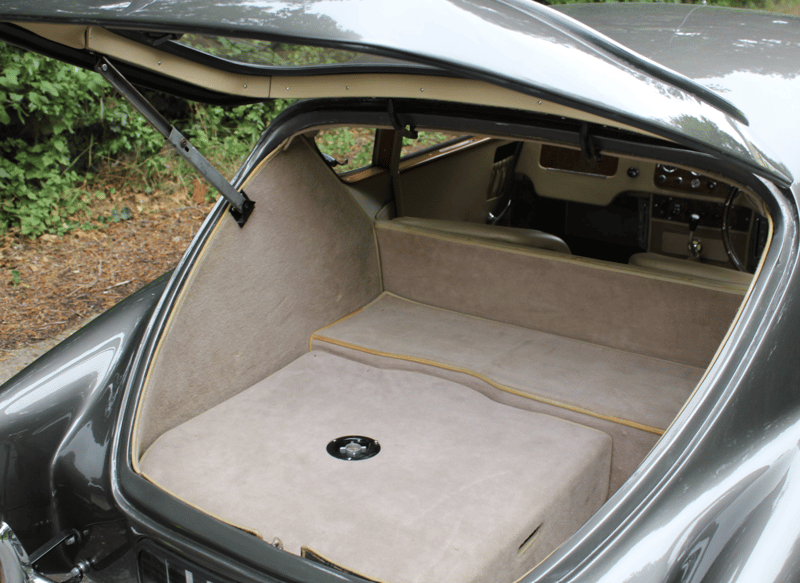 The rear of a grey AC Aceca with the one-piece tailgate raised on its stay, looking down into the carpeted luggage compartment and through to the seats and dashboard.