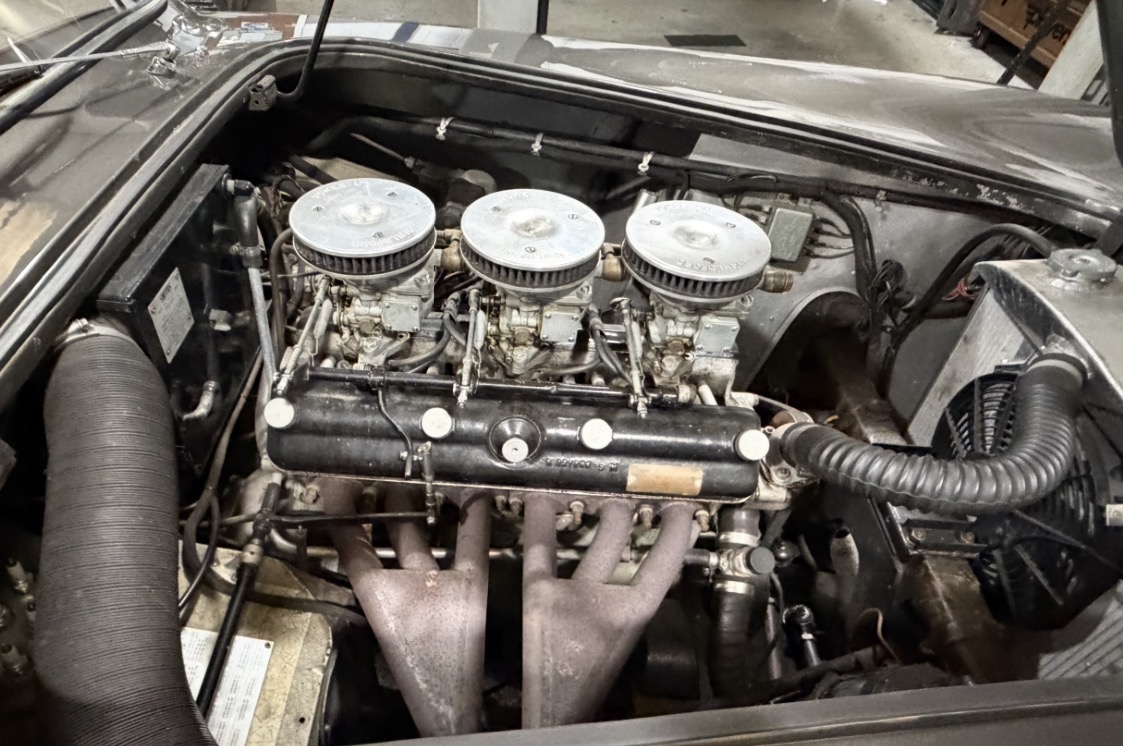 The engine bay of an AC Aceca with the bonnet raised, showing the Bristol straight-six, its black rocker cover, three downdraught Solex carburettors with polished air filters, and the alloy radiator alongside.