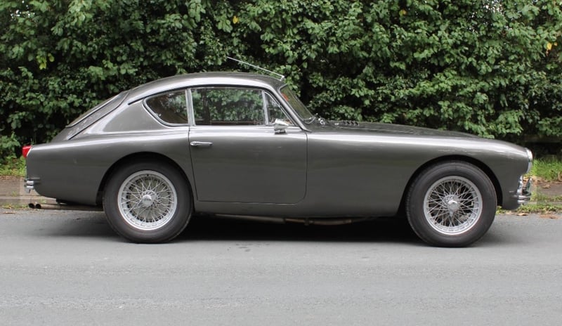 A grey AC Aceca-Bristol photographed from the nearside, parked on a road in front of a tall green hedge, showing the fastback roofline, chrome wire wheels and side exhaust.